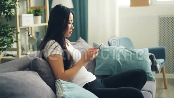 可爱的亚洲女学生正在使用现代智能手机触摸屏幕和微笑坐在沙发上独自在家青年视频的预览图