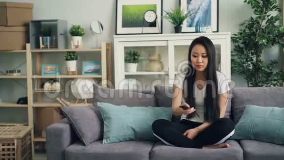 好看的亚洲女孩使用遥控器坐在沙发上在家看电视的肖像女青年休闲装视频的预览图