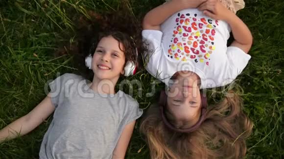 两个戴着耳机的可爱女孩躺在公园里美丽的孩子上学时代正在一起度过快乐的时光视频的预览图
