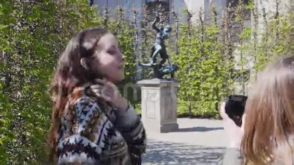 两个年轻的女人在公园里的装饰植物中散步用雕像拍照视频的预览图