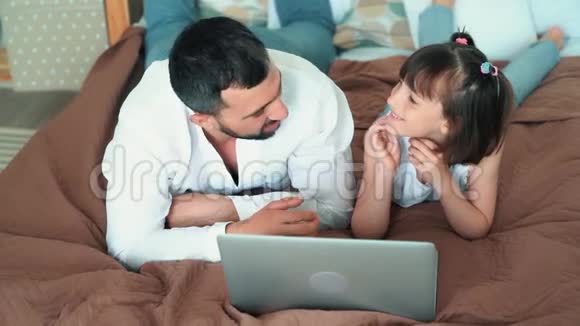 爸爸和可爱的孩子用笔记本电脑躺在床上玩网络游戏慢动作视频的预览图