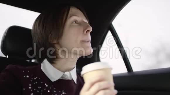 穿着白色上衣和栗色毛衣的棕色头发的漂亮女人坐在车里喝着咖啡视频的预览图