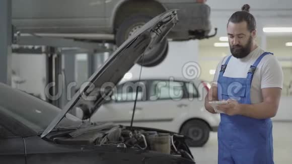 成年机械师站在汽车附近带着一个打开的引擎盖并在他的手机上验证信息穿制服的胡子男视频的预览图