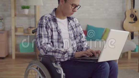 戴着时髦眼镜的年轻亚裔男子轮椅残疾人家里客厅里有一台笔记本电脑视频的预览图