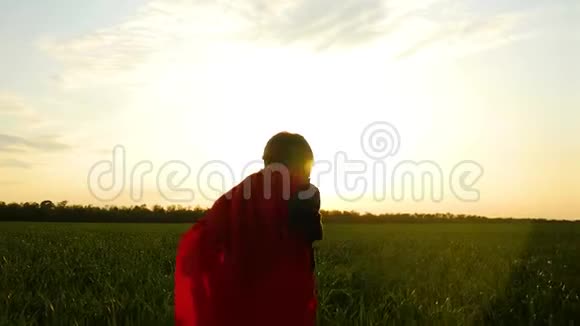 一个穿着超级英雄服装的快乐的孩子穿过绿色的草坪走向日落视频的预览图