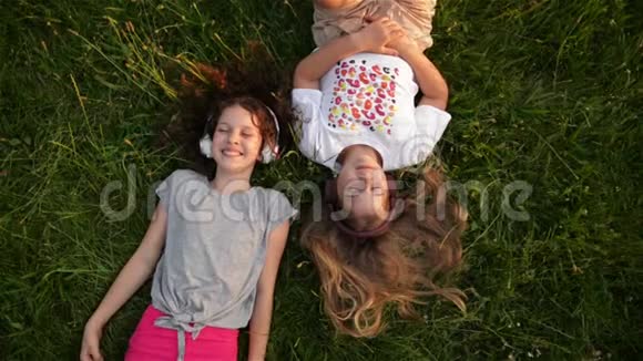两个戴着耳机的可爱女孩躺在公园里美丽的孩子上学时代正在一起度过快乐的时光视频的预览图