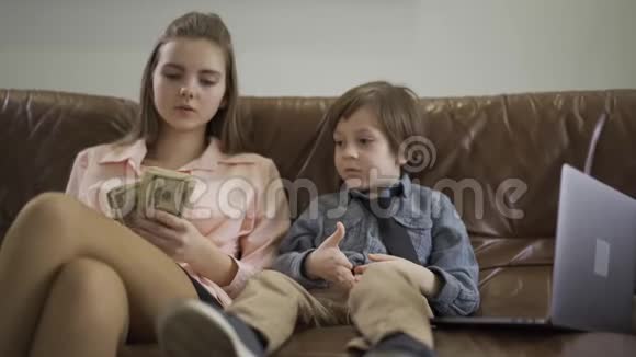 严肃的姐姐和弟弟坐在真皮沙发上笔记本电脑就在附近女孩数着钱和视频的预览图