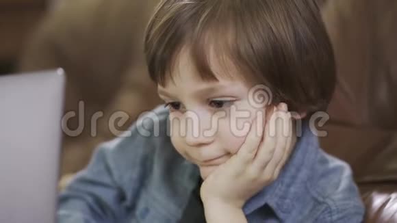 肖像可爱的小男孩穿着牛仔裤夹克坐在皮革沙发上玩着笔记本电脑开心地笑着这就是视频的预览图