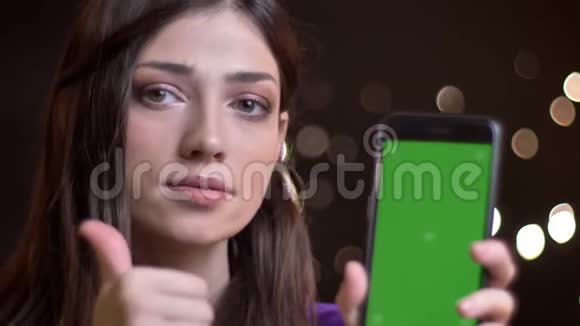 用显示绿色屏幕的手机拍摄年轻漂亮的白种人女性的特写肖像并用大拇指示意视频的预览图