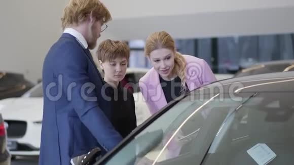 穿着粉色夹克的女售货员和站在汽车附近透过窗户往里面看的顾客讨论这辆车视频的预览图
