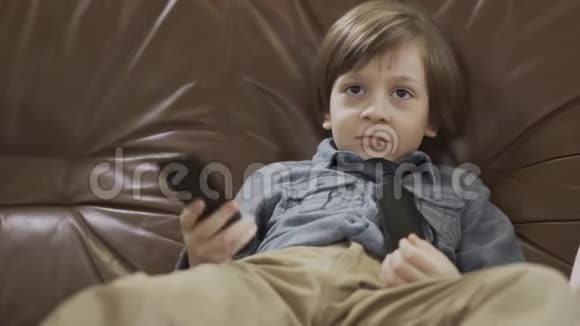 肖像可爱的小男孩坐在皮革沙发上双腿分开用遥控器在电视上更换频道可爱的儿童视频的预览图
