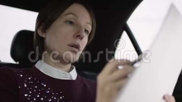 穿着白色上衣和栗色毛衣的棕色头发的漂亮女人坐在车上学习工作文件视频的预览图