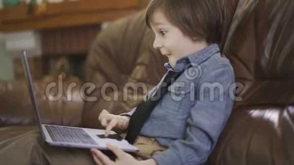 那个穿着牛仔裤夹克的小男孩坐在皮沙发上玩手提电脑看着屏幕惊讶不已视频的预览图