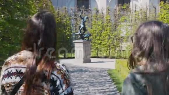 两位年轻妇女在公园里的装饰植物中散步并为雕像拍照视频的预览图
