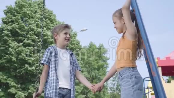 可爱的男孩和美丽的女孩长发牵着手在秋千附近说话开心地笑着一对快乐视频的预览图