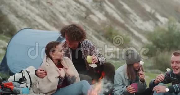 一对年轻夫妇和他们的朋友们一起在露营晚上在篝火旁吃东西视频的预览图