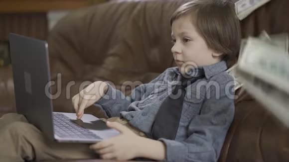 那个穿着牛仔裤夹克的小男孩坐在皮革沙发上玩笔记本电脑一美元落在沙发上孩子视频的预览图