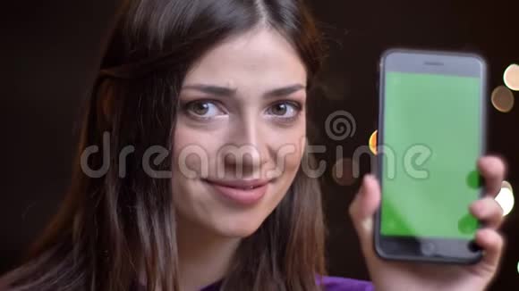 年轻迷人的白种人女性的特写肖像手持一部手机指向绿色屏幕微笑着展示视频的预览图