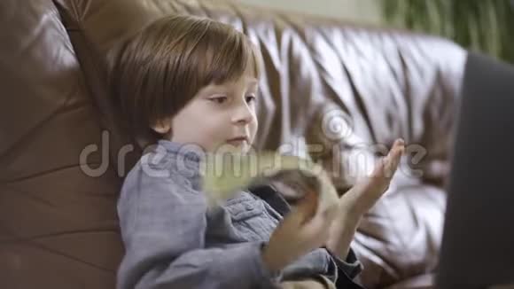 那个穿着牛仔裤夹克的小男孩坐在笔记本电脑前的皮革沙发上玩钱一捆视频的预览图