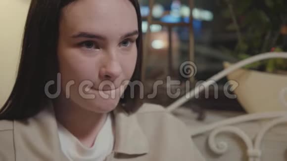 一个年轻漂亮的女孩在晚上坐在咖啡馆或餐馆的露台上沉思的肖像视频的预览图