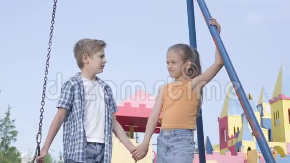可爱的男孩和美丽的女孩长发牵着手在秋千附近说话开心地笑着一对夫妇视频的预览图