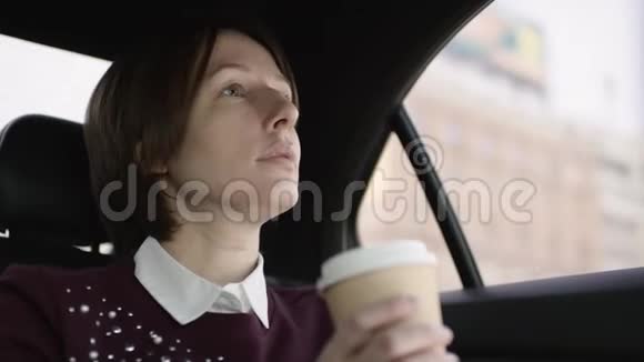 穿着白色上衣和栗色毛衣的棕色头发的漂亮女人坐在车上喝咖啡视频的预览图