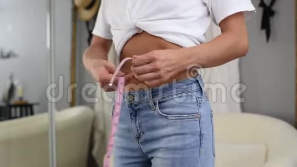 穿牛仔裤站在镜子前的幸福女人正在测量减肥后的腰围视频的预览图