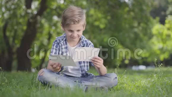 一幅可爱的可爱男孩穿着格子衬衫看着公园里的纸的画像户外夏日休闲视频的预览图