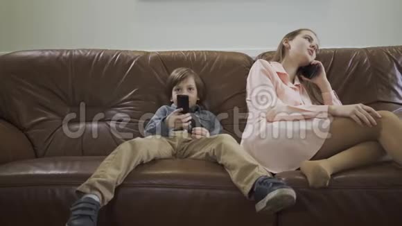 小可爱的男孩坐在皮革沙发上两腿分开用遥控器在电视上换频道而姐姐则用遥控器视频的预览图