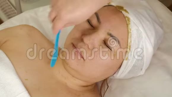 美容师从一个亚洲女人的脸上取出海藻或猕猴桃中的化妆面具的遗骸一张脸视频的预览图