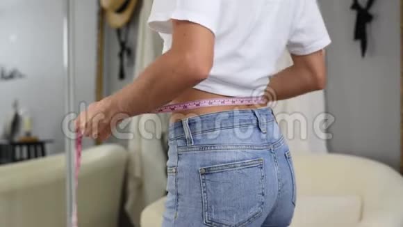 穿牛仔裤站在镜子前的幸福女人正在测量减肥后的腰围视频的预览图