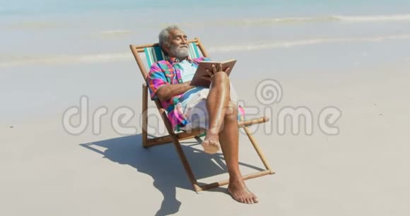 正面观看活跃的非裔美国人老人在甲板椅上放松海滩上有一本书视频的预览图