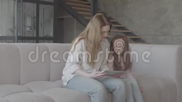 美丽的年轻母亲和她可爱的小女儿正在大房子里坐在沙发上用平板电脑微笑着视频的预览图