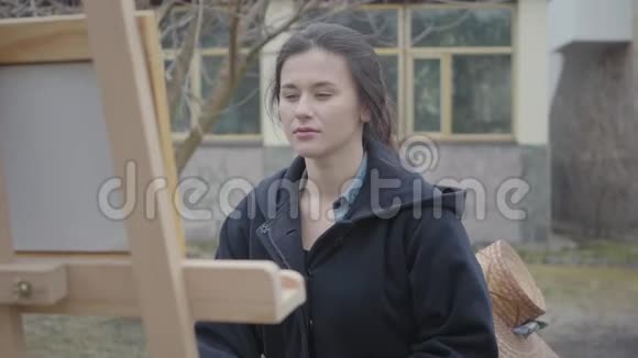 自信的年轻女孩画家坐在木制画架前环顾四周女艺术家在休闲关门中视频的预览图