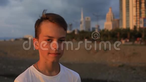 日落时分一个十几岁的男孩在城市背景下海边的肖像视频的预览图