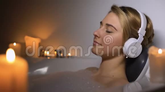 戴耳机的积极女孩躺在浴缸里用泡沫和蜡烛听音乐视频的预览图