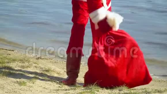 圣诞老人拿着一袋礼物站在海边圣诞老人把一个带礼物的袋子放在沙子上像腿一样视频的预览图