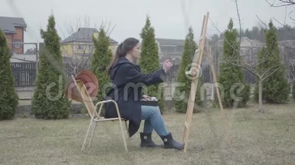 漂亮的年轻女孩画家坐在木制画架前画一幅画女艺术家在休闲结束时的花费视频的预览图