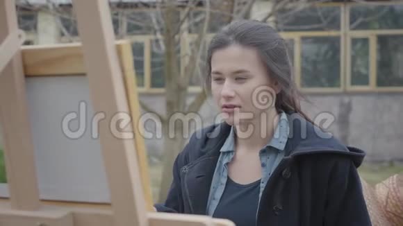肖像成功美丽的年轻女孩画家坐在木制画架前环顾四周女性艺术家视频的预览图