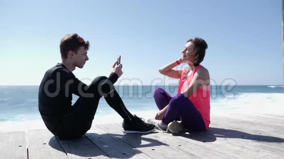 年轻的健康男子坐在海滩上用电话给他的女朋友拍照强烈的海浪拍打着岩石视频的预览图