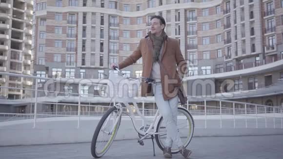 穿着棕色外套和浅蓝色牛仔裤的英俊严肃的男子骑着自行车站在城市的高楼前视频的预览图