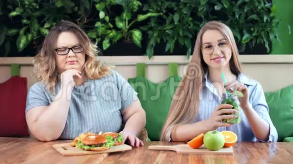 微笑苗条的女孩和悲伤肥胖的女人坐在咖啡馆里快餐和健康的饭菜视频的预览图