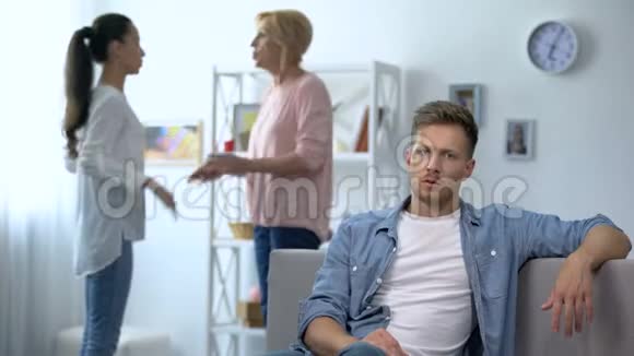 不高兴的男人坐在家里的沙发上听妻子和母亲争吵视频的预览图
