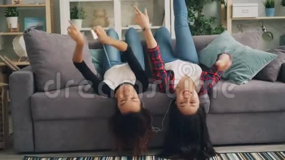 快乐的女孩朋友们正在听音乐他们用智能手机带着耳机倒着躺在沙发上跳舞视频的预览图