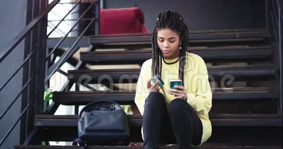 漂亮的非洲裔女士在现代办公楼梯上坐下来休息时使用智能手机和银行卡视频的预览图