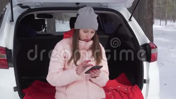 美丽的女孩坐在冬天的森林里的车里透过Instagram看着视频的预览图
