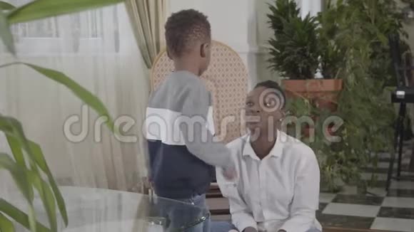 快乐美丽的非裔美国女人在舒适的客厅里和她的小有趣的儿子玩关系妈妈和视频的预览图