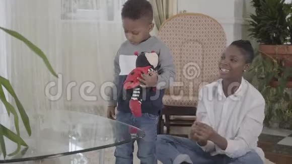 自信美丽的非裔美国女人在舒适的客厅里和她的小有趣的儿子玩妈妈视频的预览图