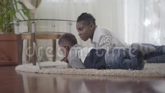 非裔美国妇女在地毯上的笔记本电脑上工作她可爱的小儿子拿着玩具来找她视频的预览图