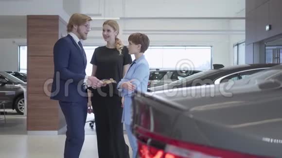 穿着西装的英俊的推销员告诉两个成功的年轻女孩关于一辆新汽车的信息视频的预览图